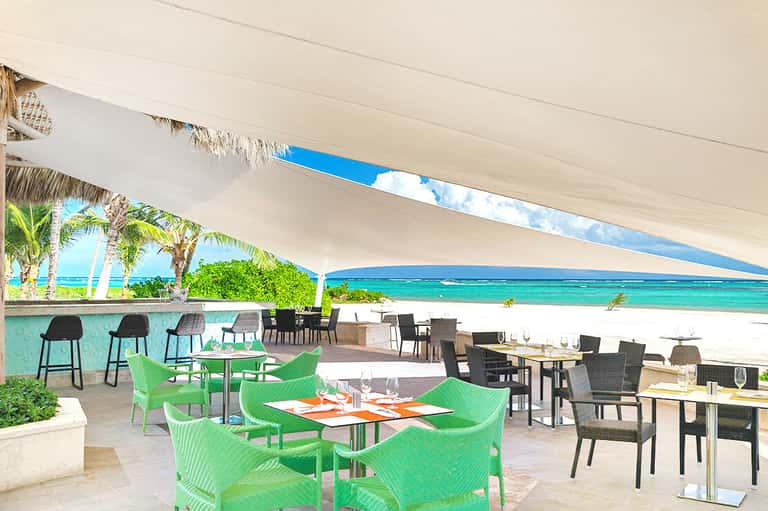 Beachfront restaurant terrace with green chairs, white shade sail, turquoise ocean view, and tropical dining ambiance
