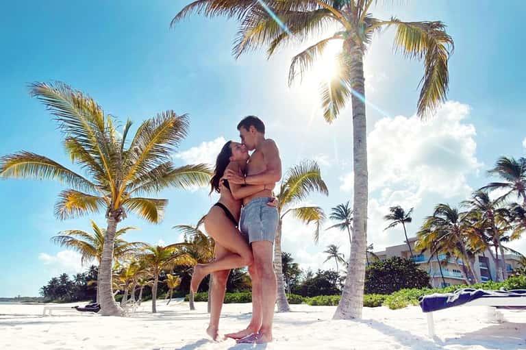 Couple embracing on a sandy beach with palm trees and resort buildings behind