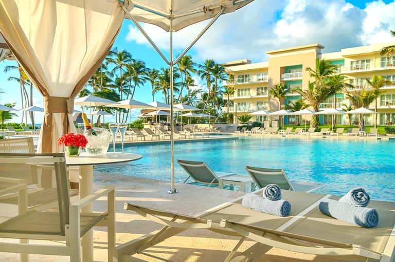 Beachfront resort pool area with palm trees, cabanas, lounge chairs, and tropical landscaping.