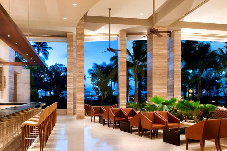 Luxury resort lounge at dusk with travertine columns, ocean views, palm trees, and burnt orange seating