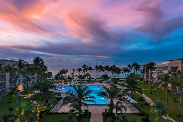 Aerial view of beachfront resort at dusk with illuminated pool, palm trees, and ocean view