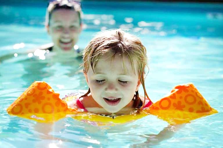 Young child swimming in bright blue pool water wearing orange flotation wings, another child visible in background