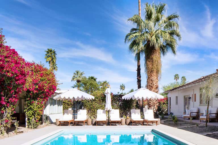Resort courtyard with turquoise pool, white umbrellas, lounge chairs, pink flowering plants, and palm trees