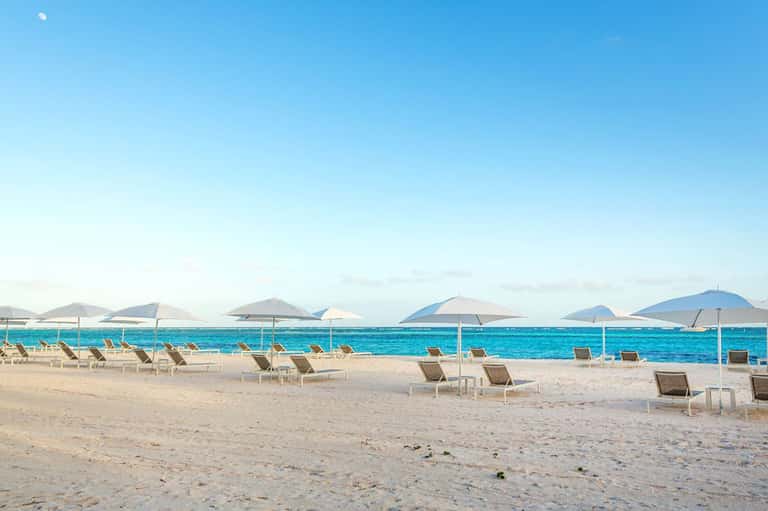 Wide sandy beach with lounge chairs, white umbrellas, and turquoise ocean under blue sky