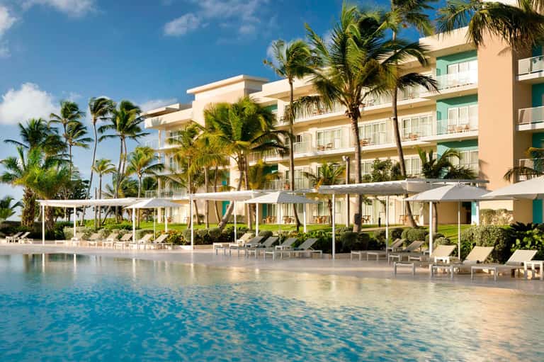 Beachfront hotel with palm trees, sandy beach access, lounge chairs with umbrellas, and colorful building facade