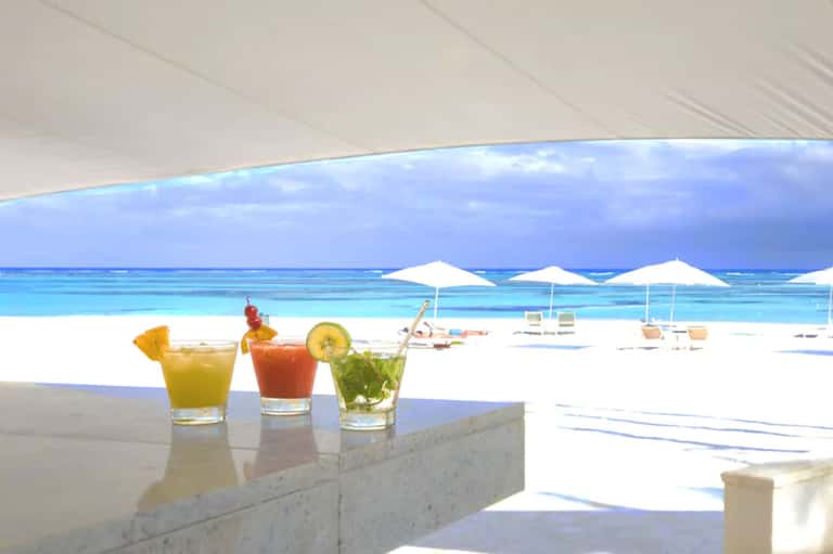 Three tropical cocktails on marble counter with beach, umbrellas, and turquoise water in background
