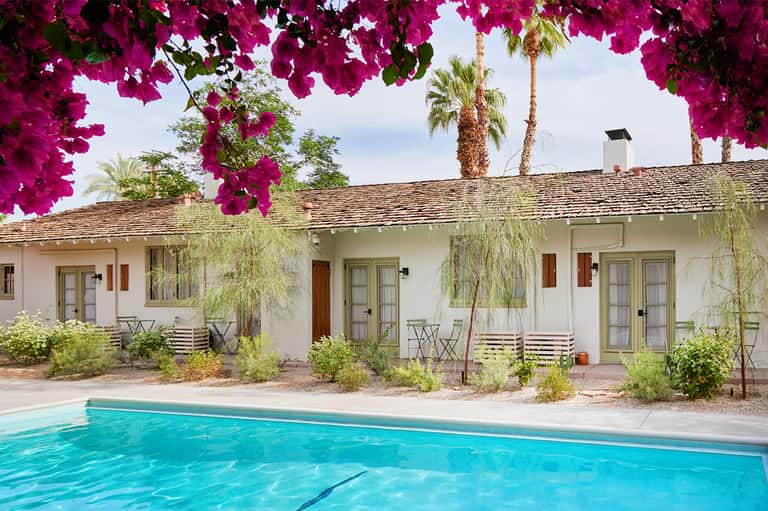White mid-century resort building with magenta bougainvillea overhead, turquoise pool in foreground, palm trees