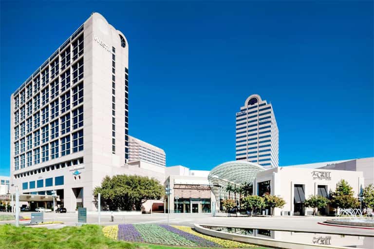 Modern hotel exterior with white high-rise towers and contemporary plaza featuring landscaped gardens and reflecting pool