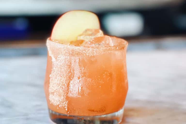 Coral-colored cocktail with sugar rim and apple slice garnish by the poolside