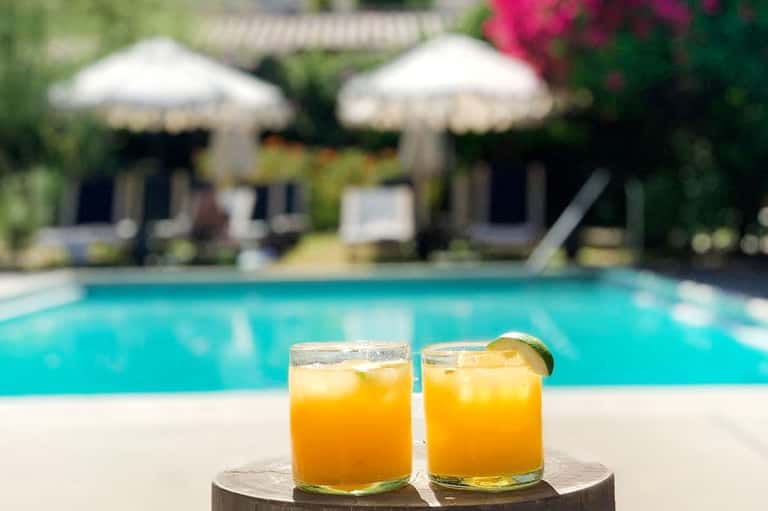 Two tropical cocktails by poolside with umbrellas, lounge chairs, and beach cabanas in background