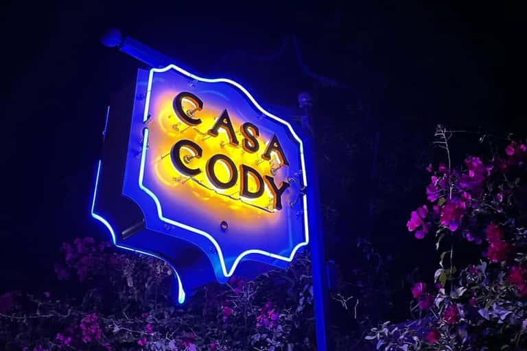 Casa Cody neon sign glowing blue and yellow at night surrounded by pink flowers