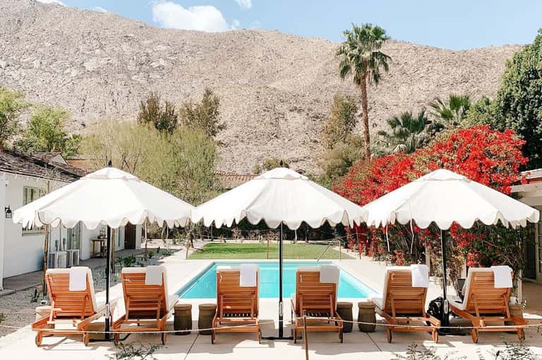Desert resort pool with white umbrellas, wooden loungers, and red flowering vines on mountain backdrop