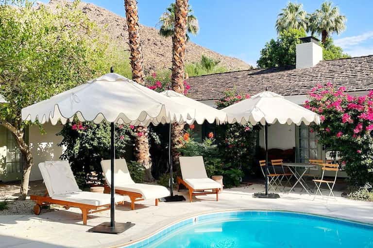 Poolside lounge area with white umbrellas, palm trees, pink flowers, and mountain landscape