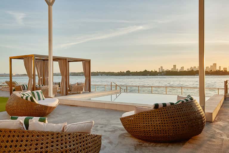 Waterfront lounge with woven pod chairs, white awnings, and city skyline view across water