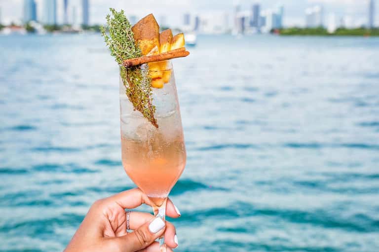 Tropical cocktail with fresh herbs and gold leaf garnish held over blue ocean water with city skyline backdrop