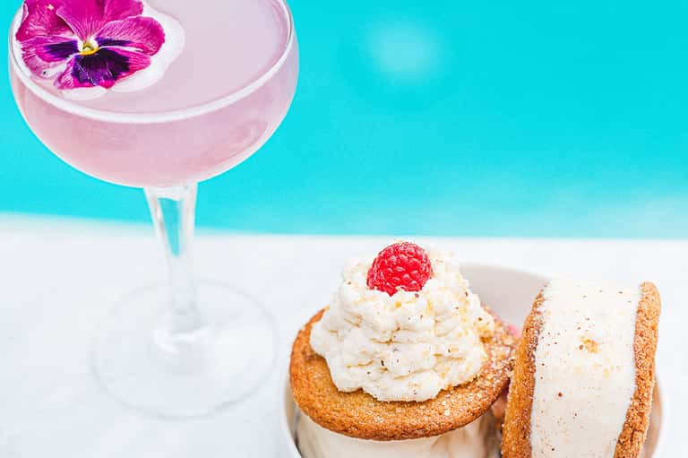 Pink cocktail with flower garnish and desserts on white sand beach with turquoise water