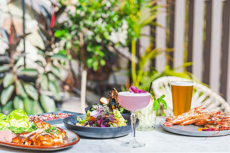 Upscale outdoor dining table with colorful cocktail, gourmet seafood, fresh salad, and lush greenery background