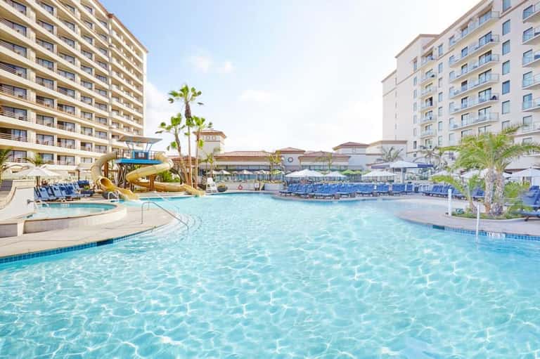 Large outdoor resort pool with water slide, palm trees, and multi-story hotel buildings under clear sky