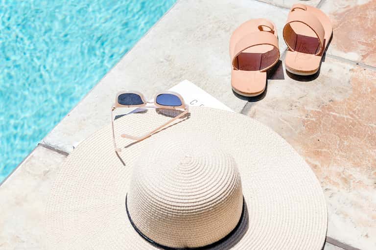 Pool deck with white wide-brimmed hat, sunglasses, and peach sandals on concrete