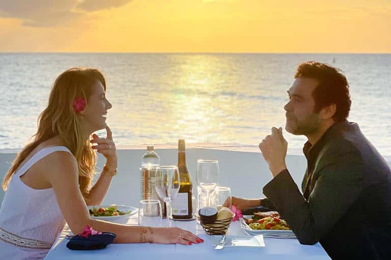 Couple dining at sunset table overlooking ocean with wine, champagne, and tropical flower decorations