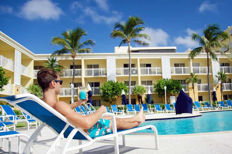 Man relaxing in lounge chair by pool at yellow and white resort hotel with palm trees