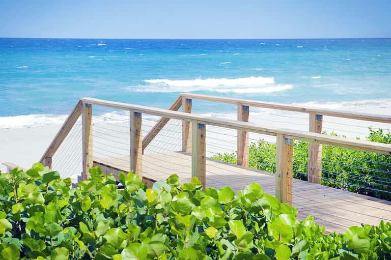 Wooden boardwalk with cable railing overlooking pristine white sand beach, ocean waves, and coastal vegetation