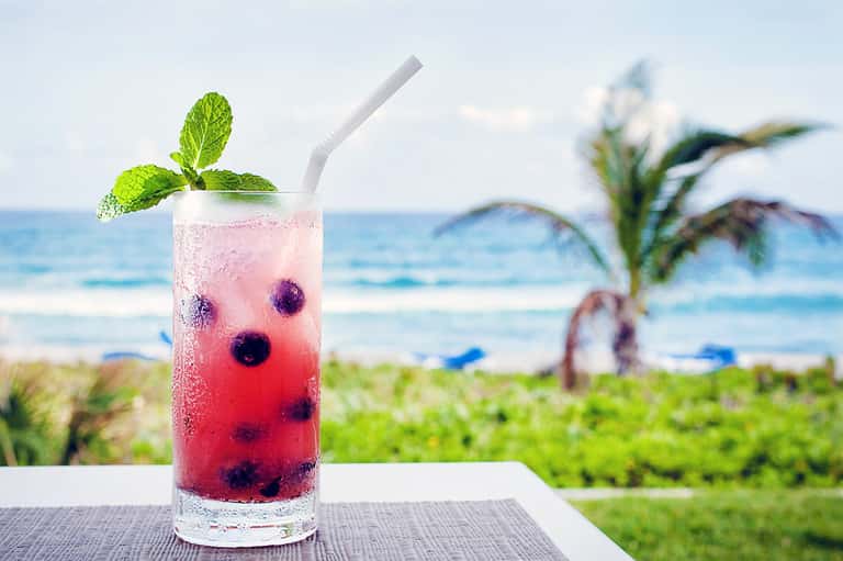 Tropical cocktail with berries and mint garnish on oceanside table with palm trees and beach
