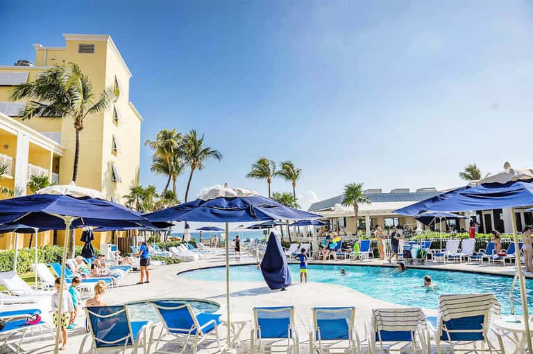 Beachfront resort pool area with yellow building, blue umbrellas, lounge chairs, and palm trees