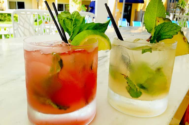 Two colorful mojito-style drinks with fresh mint, lime, and ice on outdoor white table