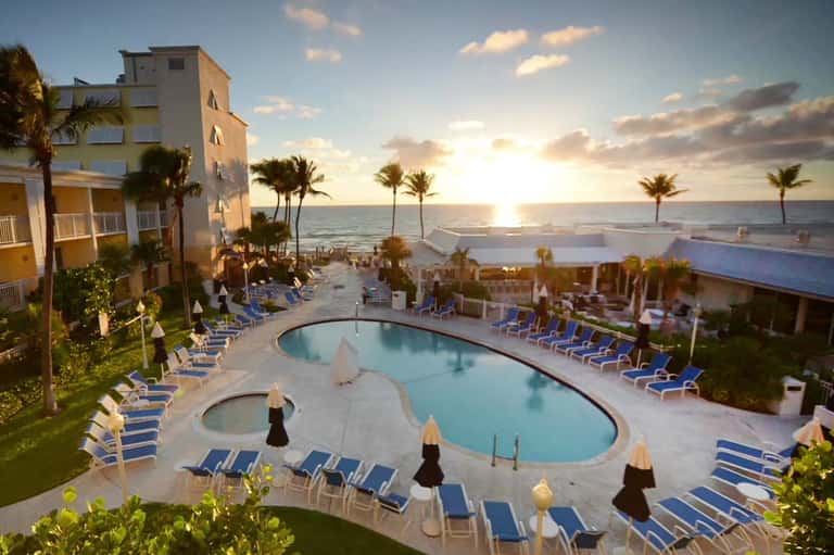 Aerial view of curved resort pool with lounge chairs, ocean, and palm trees at sunset