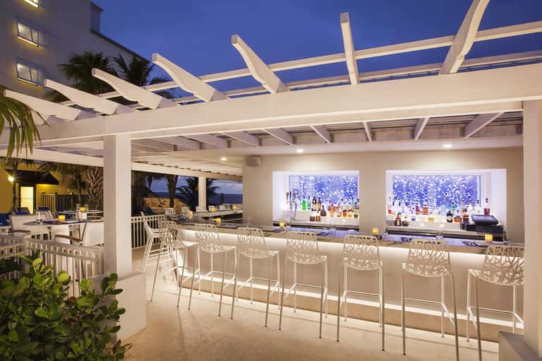 Modern beachside bar with white pergola, illuminated shelving, ocean view, and elegant seating