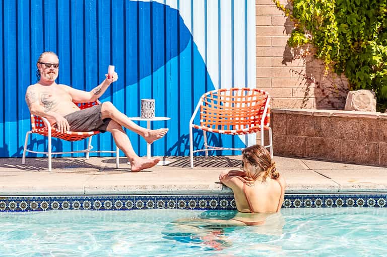 Man relaxing poolside in chair while woman swims in bright turquoise pool with blue wall mural