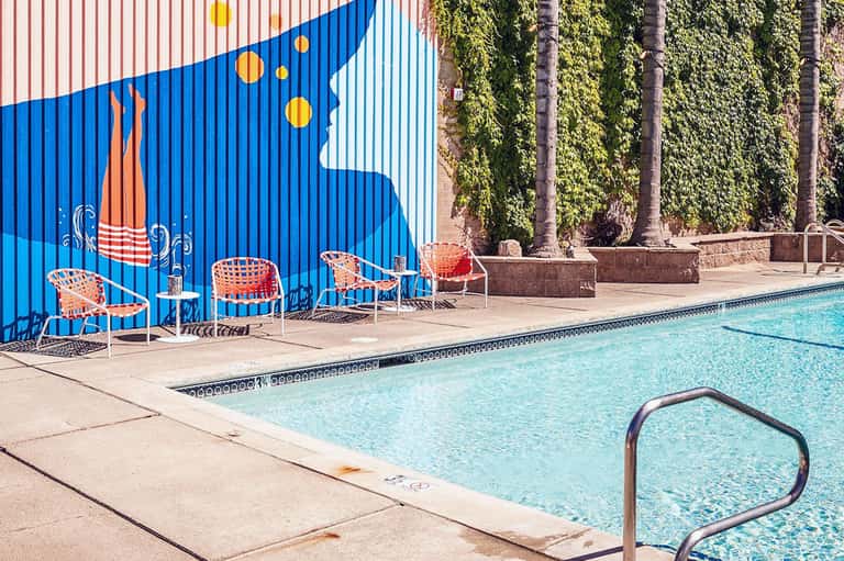Modern poolside area with bright blue mural, orange chairs, and ivy-covered walls