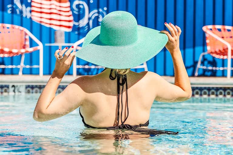 Person in pool wearing wide-brimmed green hat, holding the brim with both hands while in shallow water