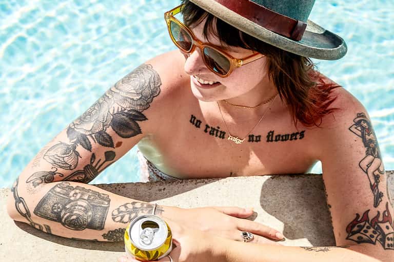 Tattooed woman in sunglasses and hat relaxing by the pool with a beverage