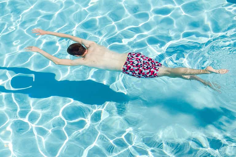 Young boy swimming backstroke in clear blue pool with colorful geometric patterned swim shorts