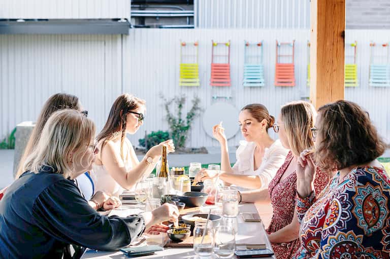 Group of women dining outdoors at long table with colorful fitness equipment on white building