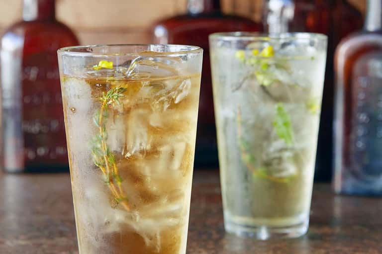 Two refreshing cocktails with herbs and ice on bar counter with bottles in soft-focus background