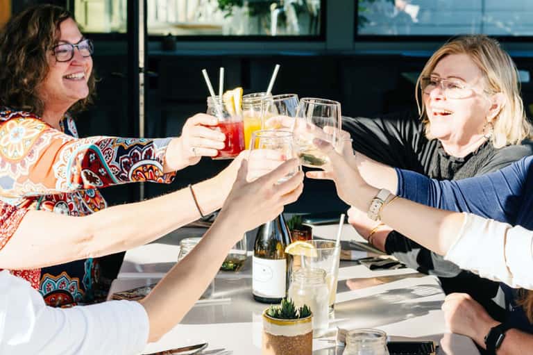 Two women toasting with colorful drinks at an outdoor dining table with ocean view in background