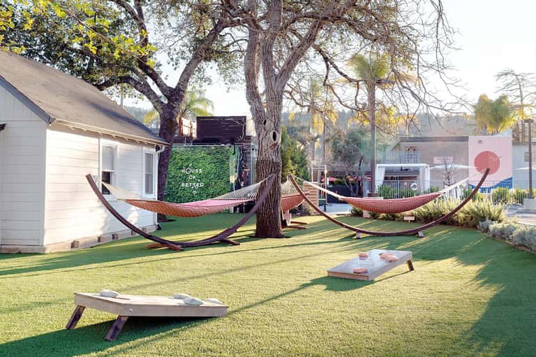 Outdoor lawn with hammocks suspended between trees, cornhole boards, and modern white buildings in background