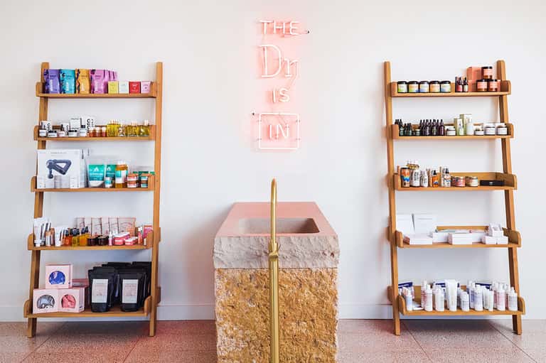 Spa retail display with wooden shelves holding skincare products and pink neon sign reading 'The Day is in'