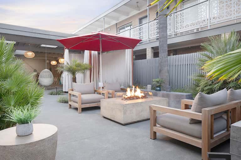 Modern courtyard with fire pit, wooden lounge chairs, red umbrella, potted plants, and contemporary architecture