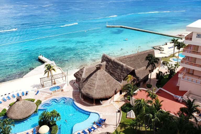 Beachfront resort with turquoise ocean, thatched-roof structures, swimming pool, and lounging chairs