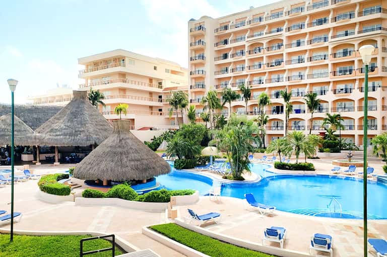 Resort pool complex with thatched roof structure, blue water, palm trees, and multi-story peach hotel building