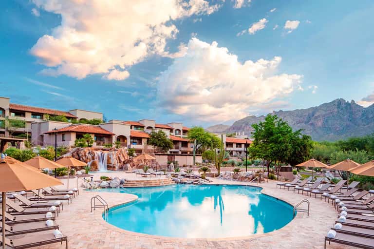 Resort pool with waterfall feature, desert mountain backdrop, terracotta buildings, and umbrella seating area