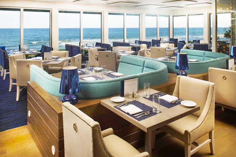Upscale dining room with curved turquoise seating, ocean views through floor-to-ceiling windows, and elegant coastal décor