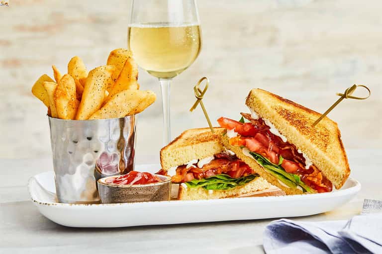 Gourmet BLT sandwich with fries and white wine on white plate