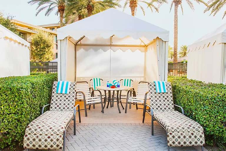 Luxury cabana lounge area with striped chairs, round table, and white canopy tent surrounded by green hedges and palm trees