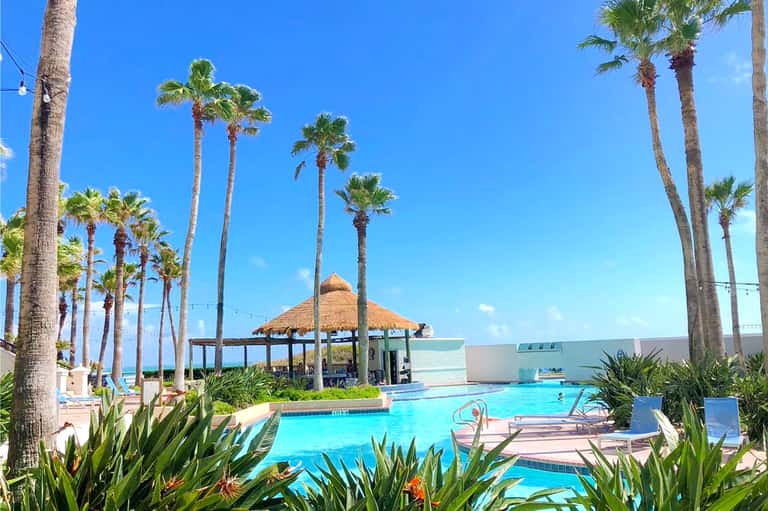 Resort pool area with thatched cabana, palm trees, and turquoise water under clear blue sky