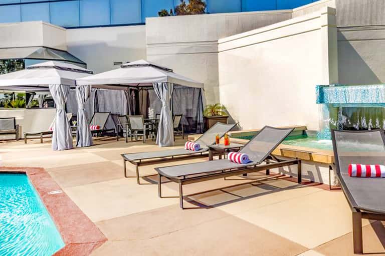 Luxury resort pool deck with lounge chairs, umbrellas, cabanas, and modern white building under clear blue sky